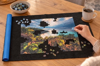 Tapis à puzzle gris déroulé sur une table avec un puzzle en cours, tasse de café et pièce tenue au-dessus